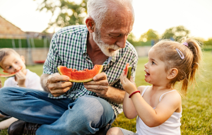 A fit and healthy senior man over 60 eating watermelon on the grass with his grandchildren – this is these are the real-life fitness goals we want over 60